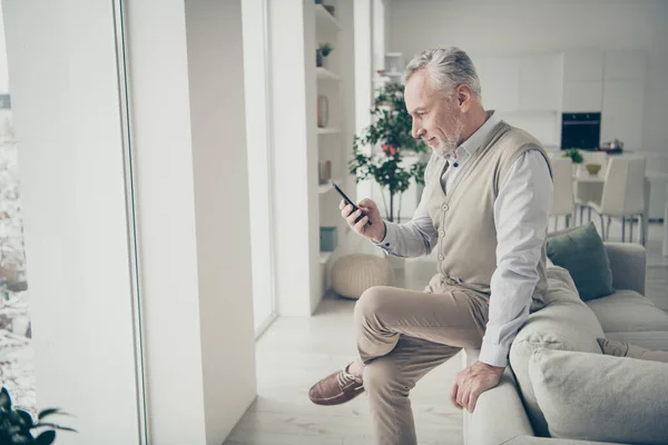 Foto de homem de cabelos brancos idosos segurando telefone mão procurando notícias sentado sofá dentro de casa — Fotografia de Stock