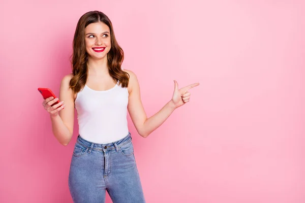 Портрет позитивної веселої дівчини використовує індекс точок смартфона палець копіяпростору пропоную вибрати рекламу одягати білі джинси з баком на верхній частині джинси ізольовані на рожевому кольорі — стокове фото
