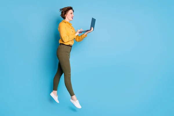 Foto de perfil de tamanho completo de bela senhora saltando alto segurando navegação notebook workaholic usar camisa amarela calças verdes sapatos isolados fundo de cor azul brilhante — Fotografia de Stock