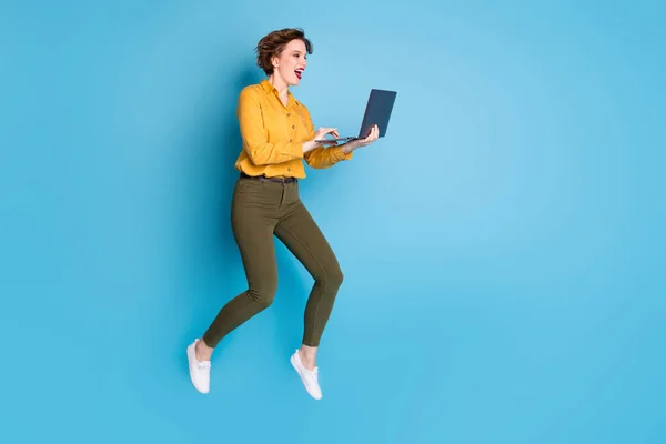 Full length profile photo of pretty lady jumping high up holding browsing notebook workaholic wear yellow shirt πράσινο παντελόνι παπούτσια απομονωμένο φωτεινό μπλε χρώμα φόντο — Φωτογραφία Αρχείου