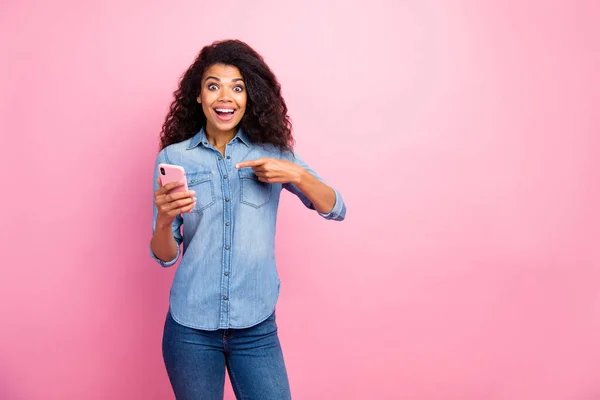 Vypadat černě první den. Závislý překvapený afro americká dívka použít smartphone point ukazováček dojem sociální sítě zprávy oblečení džíny košile izolované přes růžové barvy pozadí — Stock fotografie