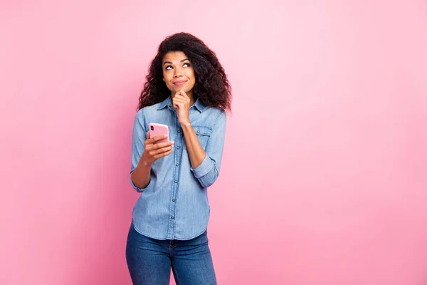 Retrato de una bloguera afro-americana adicta a las redes sociales inspirada usa el teléfono celular piensa qué pensamientos planean qué artículo en el blog usa una camisa vaquera aislada sobre un fondo de color rosa — Foto de Stock