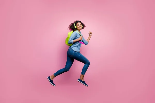 Volledige lengte profiel zijkant foto van vrolijke afro-Amerikaanse meisje college student jump run haast lezing luisteren muziek headset wear denim jeans shirt tas reizen geïsoleerde roze kleur achtergrond — Stockfoto