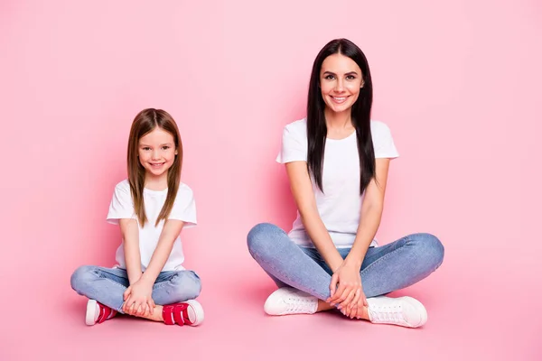 Volledige lengte foto van aantrekkelijke jonge moeder kleine dochter goed humeur beste vrienden zit vloer gekruiste benen dragen casual witte t-shirts jeans geïsoleerde pastel roze kleur achtergrond — Stockfoto