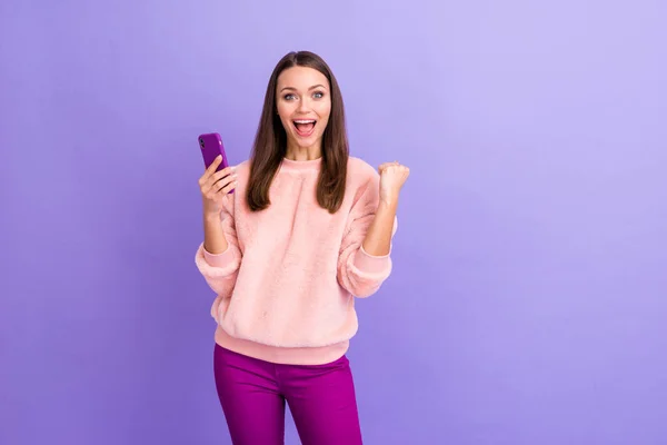 Foto de una bloguera bastante adicta que sostiene el teléfono celebrando un gran número de nuevos seguidores que usan pantalones de suéter esponjosos de piel casual aislados de color púrpura de fondo — Foto de Stock