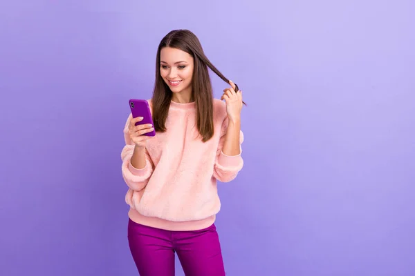 Foto de blogueiro muito viciado senhora segurando telefone mensagens de texto amigos brincando com longos cachos desgaste casual pele macia pulôver calças isoladas cor roxa fundo — Fotografia de Stock