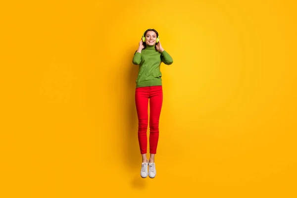 Full length φωτογραφία της όμορφης κυρίας άλμα ψηλά κρατώντας τα αυτιά με δροσερά σύγχρονα ακουστικά ακούγοντας μουσική φορούν casual πράσινο jumper κόκκινο παντελόνι απομονωμένο κίτρινο χρώμα φόντο — Φωτογραφία Αρχείου