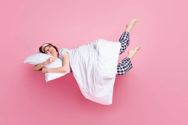 Фотография в полный рост веселой дамы, удовлетворенной утренним сном, лежащей на кровати, обнимающей подушку одеяло, поднимающей ноги вверх, надевающей маску белую футболку и пижамные брюки барочного цвета на розовом фоне — стоковое фото