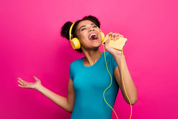 Foto av ganska funky mörk hud dam hålla telefon lyssna musik moderna hörlurar sjunga smart telefon som mikrofon bära blå casual t-shirt isolerad ljus rosa färg bakgrund — Stockfoto