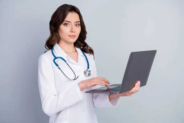 Портрет досвідченої дівчини-інфекціоністки використовує ноутбук, готовий для лікування ковідо-19 пневмонії, одягають біле пальто ізольоване на сіро-кольоровому фоні — стокове фото
