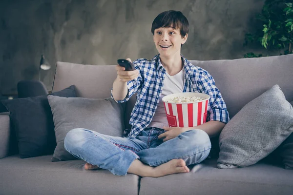 Photo of teenager school boy sit comfy couch stay home quarantine time watch movie show eat popcorn good mood changing channels legs crossed living room indoors