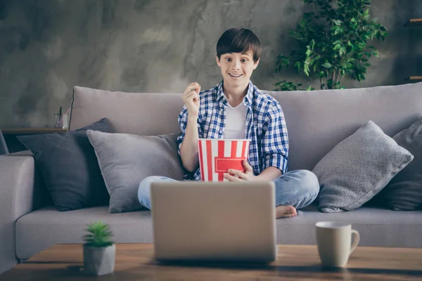 Фото подростка, сидящего на удобном диване, сидящего дома во время карантина и смотрящего онлайн-фильм о блокноте, который ест попкорн. — стоковое фото
