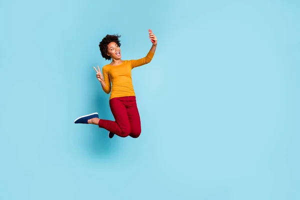 Full length body size view of nice attractive cheerful wavy-haired girl jumping taking selfie on cell phone smartphone showing v-sign isolated on bright vivivid shine vibrant blue color background — стоковое фото