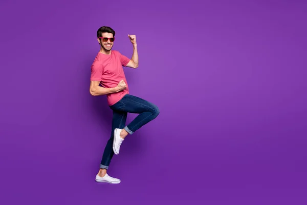 Full längd foto av snygg attraktiv kille strålande leende glädje fest började höja nävar bära solglasögon casual rosa t-shirt jeans isolerad lila färg bakgrund — Stockfoto