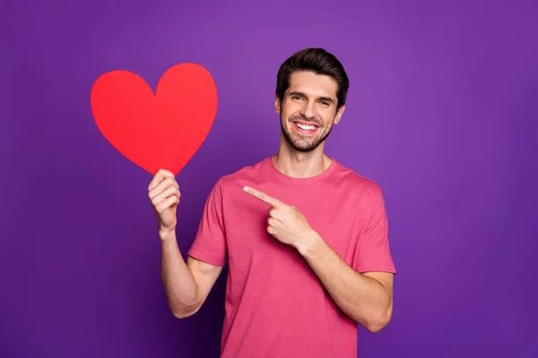 Look i have paper card heart. Portrait of positive cheerful guy hold red 14-february gift point index finger indicate ear style stylish trendy outfit isolated over shine color background — Stock Photo, Image