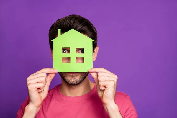 漂亮男人拿着绿纸房子房屋建筑地产代理透过小窗看隐藏着的休闲装粉红T恤孤立的紫色背景 — 图库照片