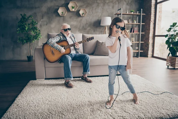 Photo of grandpa play guitar little pretty granddaughter hold mic singing song spend time rejoicing cool style trendy sun glasses stay home quarantine modern living room indoors