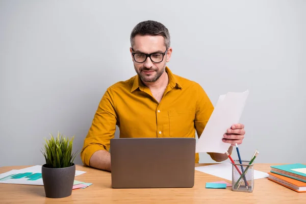 Gericht marketeer ceo man zitten bureau werk thuis covid-19 quarantaine analyseren start-up ontwikkeling innovatie grafiek gelezen laptop hold papier dragen geel shirt geïsoleerde grijze kleur achtergrond — Stockfoto