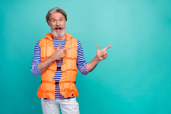 Photo of handsome shocked aged seaman directing fingers empty space lifeguard sea voyage ad banner wear striped sailor shirt shorts orange life vest isolated teal color background