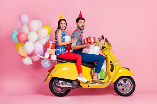 Tam boy fotoğraf çılgın pozitif enerjik iki kişi sarı retro motorlu bisiklet kullanıyor hediye kutuları var uçan balonlar var külah gömlekli kırmızı pantolon, izole pembe arka plan. — Stok fotoğraf