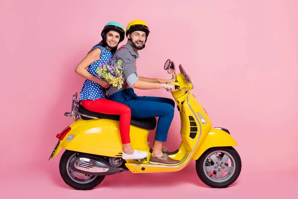 Foto lateral de perfil de cuerpo completo de motociclistas positivos dos conductores de personas conducen scooter rápido amarillo disfrutar de la aventura de campo rural mantenga flores silvestres aisladas sobre fondo de color rosa —  Fotos de Stock
