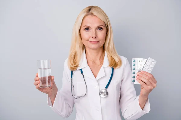 Portrait of experienced retired pensioner infection specialist woman consult corona virus patient hold pills glass water wear stethoscope isolated over gray color background — Stock Photo, Image