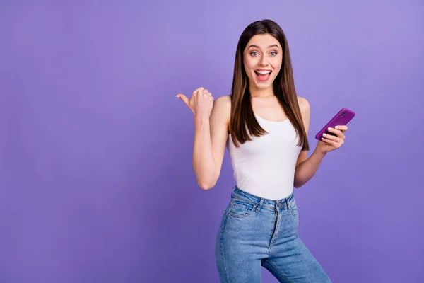 Retrato de menina positiva surpreso uso busca smartphone mídia social notícias indicam anúncios promo impressionado ponto polegar copyspace desgaste singlet denim isolado sobre fundo cor violeta — Fotografia de Stock