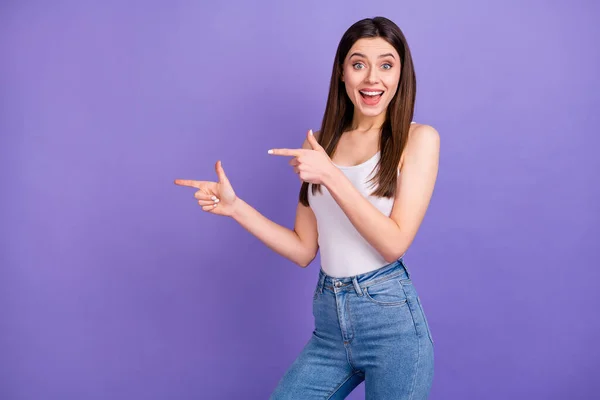 Photo of pretty lady cheerful person direct fingers side empty space advising novelty product banner low prices wear white tank-top jeans isolated pastel purple color background
