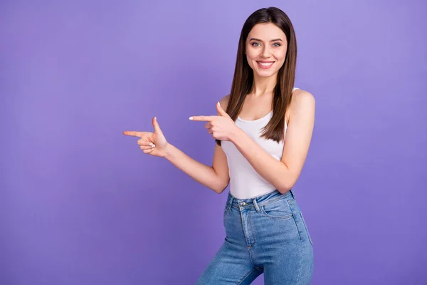 Photo of pretty lady cheerful self-confident person direct fingers side empty space advising novelty product banner wear white tank-top jeans isolated pastel purple color background