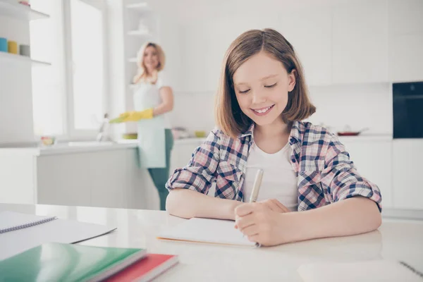 奇妙な2人の娘の母親の関係の概念。小さな子供の女の子は、テーブルに座る書き込み宿題ノートブック母でエプロンきれいな洗浄料理は、屋内キッチンハウスで隔離をお楽しみください — ストック写真