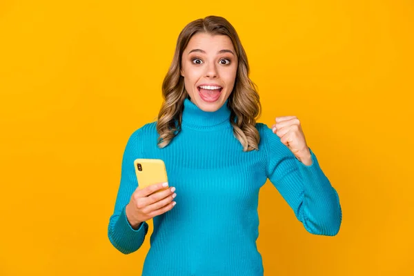 Foto van aantrekkelijke golvende gekke dame blogger houden telefoon stijging vuist vieren een miljoen volgelingen dragen casual blauwe coltrui geïsoleerde levendige gele kleur achtergrond — Stockfoto