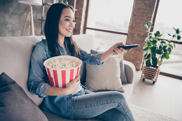 Profil side foto af charmerende oprigtig pige sidde divan hold store pop majs boks tage fjernbetjening søgning tv interessant film i lejligheden værelse - Stock-foto