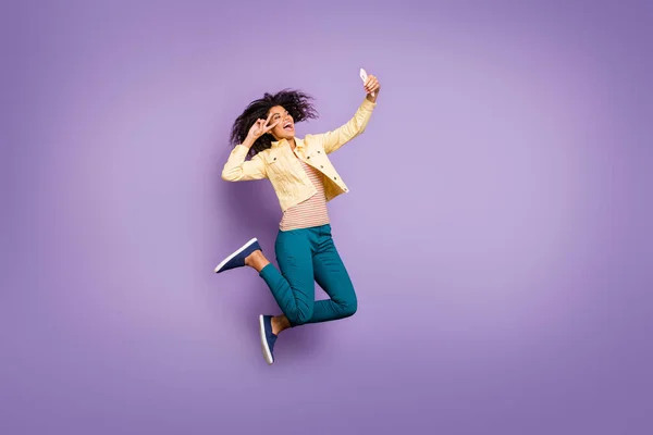 Turned full length body size photo of cheerful crazy excited girl in pants trousers showing v-sign taking selfie jumping isolated curly wavy brown hair isolated pastel violet color background