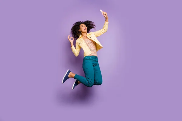 Full length body size turned photo of stylish trendy curly wavy brown hair fashion girl showing ok sign taking selfie jumping in yellow jacket pants trousers isolated pastel color violet background