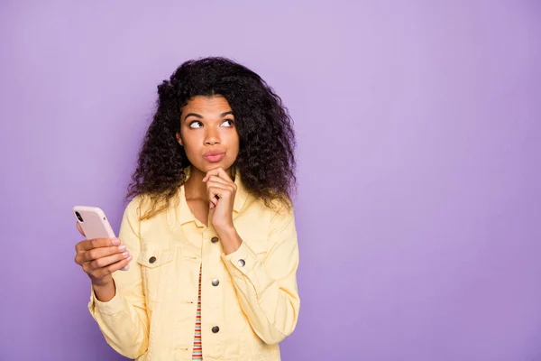Photo of curly brown haired attractive bewildered woman remembering details of chat while browsing through telephone near empty space isolated pastel color background violet — Stock Photo, Image