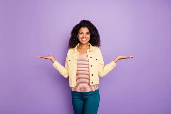 Portrait of confident afro american girl promoter hold hand present object products wear casual style clothing blue pants trousers isolated over purple color background — Stock Photo, Image