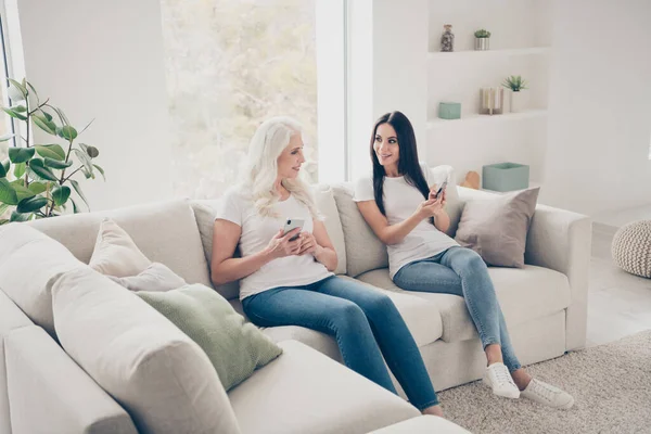 Портрет двух симпатичных привлекательных веселых женщин, сидящих на диване с помощью цифрового устройства веб-приложение проводит выходные в белом светлом интерьере квартиры — стоковое фото