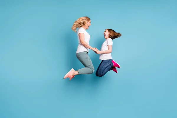 Full längd profil foto av rolig mamma dam lilla dotter tillbringa tid tillsammans hoppa högt upp hålla händerna glädje bära casual vit s-shirts isolerad blå färg bakgrund — Stockfoto