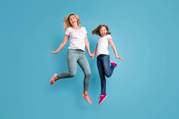 Komik anne ve küçük kızın uzun bir fotoğrafı yüksekte zıplayarak el ele tutuşarak iyi bir ruh hali içinde günlük beyaz tişörtler giyerek izole edilmiş mavi arka plan. — Stok fotoğraf