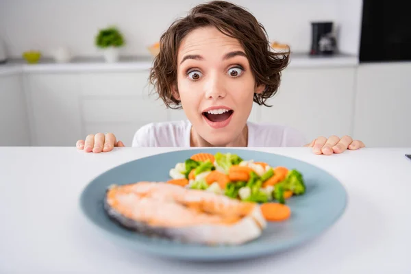 Close up foto di divertente casalinga signora astuzia occhi affamati ingannevoli guardare da sotto tavolo pronto da mangiare grigliato filetto di trota bistecca porzione guarnire cucina moderna al coperto — Foto Stock