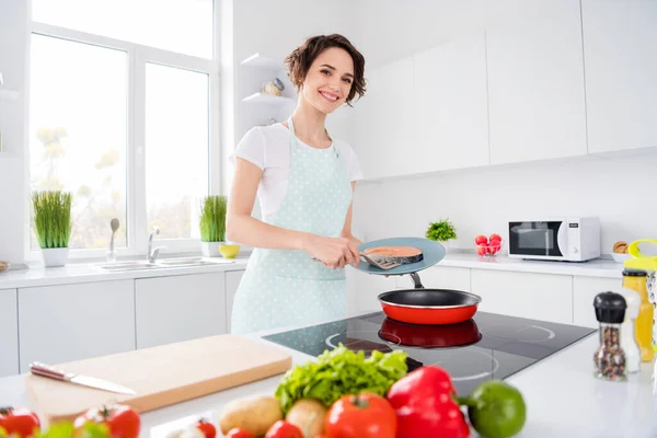 美しい陽気な主婦の女性の写真は、新鮮な生のサーモンフィレステーキをフライングパンに置きます食事の朝の料理の摩耗エプロンTシャツスタンドモダンなキッチン屋内 — ストック写真