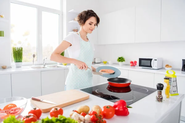 Foto van mooie huisvrouw dame zet verse rauwe zalm filet biefstuk op vliegende pan houden dieet ochtend koken dragen schort t-shirt staan moderne keuken binnen — Stockfoto