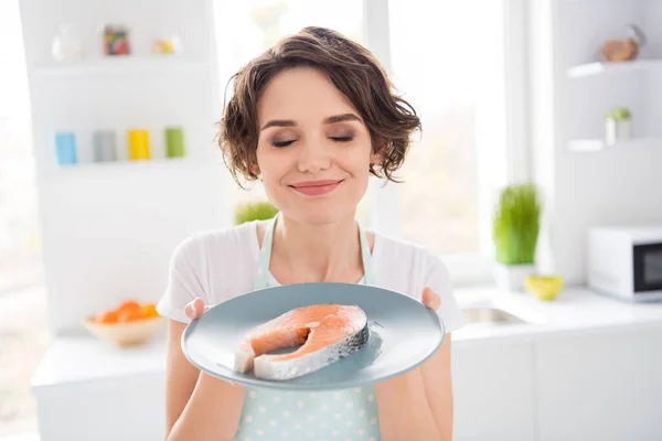 De cerca la foto de la hermosa ama de casa disfrutar de agradable olor a filete de salmón crudo fresco sabrosa dieta desayuno ojos cerrados hambre usar delantal camiseta pie cocina moderna en el interior — Foto de Stock