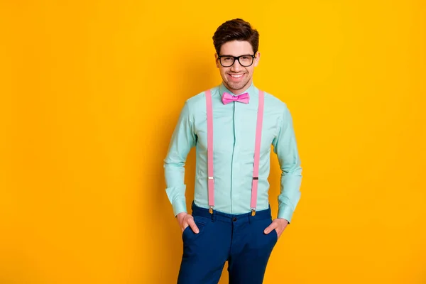 Photo of handsome cool clothes guy boyfriend standing self-confidently hands pockets beaming smile wear specs shirt suspenders bow tie trousers isolated yellow color background