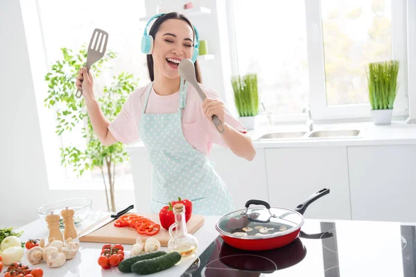 Foto van vrolijke aziatische etniciteit huisvrouw snijden salade koken lekker ontbijt luisteren koele draadloze hoofdtelefoon zingen tafel lepel favoriete lied stand moderne keuken binnen — Stockfoto