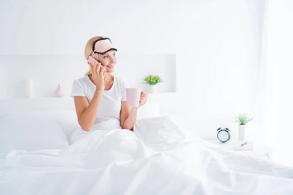 Retrato de ella ella ella agradable atractivo alegre alegre mujer mayor beber leche caliente sentado en la cama hablando por teléfono en la moderna luz blanca habitación interior apartamento en el interior — Foto de Stock