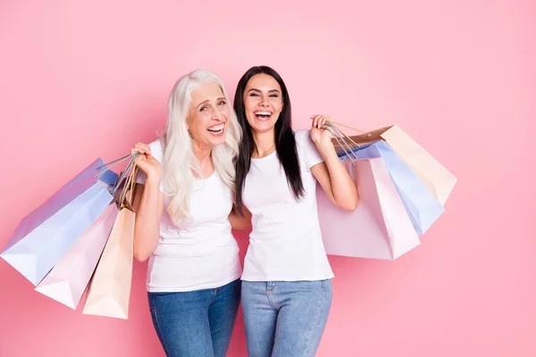 Фото смешного возраста и молодых двух дам родственников держать много магазинов пакеты ходить по магазинам вместе радуясь носить белые футболки джинсы изолированные пастельно-розовый цвет фона — стоковое фото