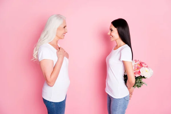 Profilo foto di allegra madre invecchiata giovani donne figlia trascorrere del tempo insieme tenendo mazzo di fiori freschi dietro la schiena indossare bianco t-shirt jeans isolato pastello colore rosa sfondo — Foto Stock