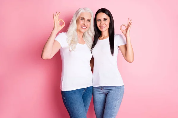 Фото довольно пожилой матери юные леди, обнимающие руки, показывающие хорошо символы, советующие новинки носить белые футболки джинсы изолированные пастельно-розовый цвет фона — стоковое фото