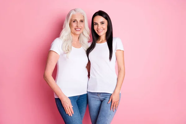 Foto de alegre bastante anciana madre joven hija damas generación abrazos pasar tiempo juntos usar blanco camisetas jeans aislado pastel rosa color fondo — Foto de Stock